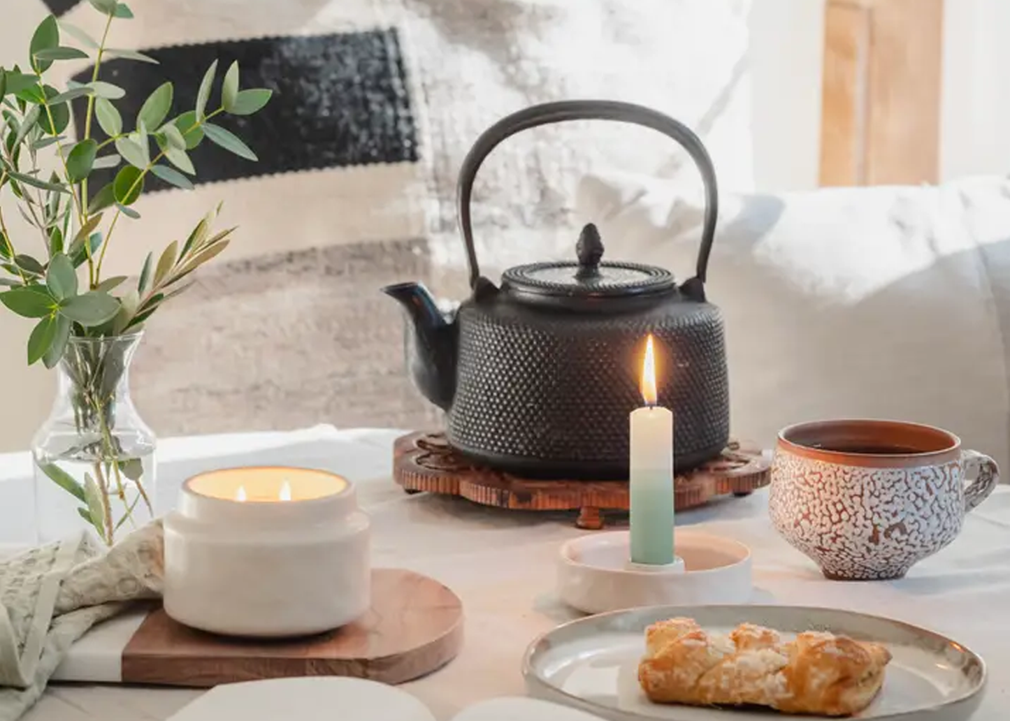 
                  
                    Tea set with teapot, cup, and candles on a table with a cozy background.
                  
                