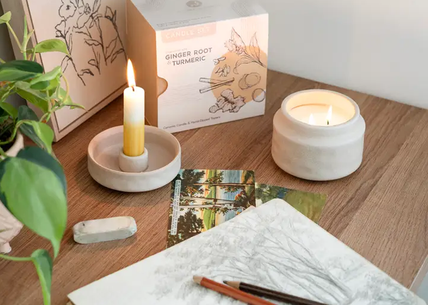 
                  
                    Two lit candles on a wooden surface with a plant and books in the background
                  
                
