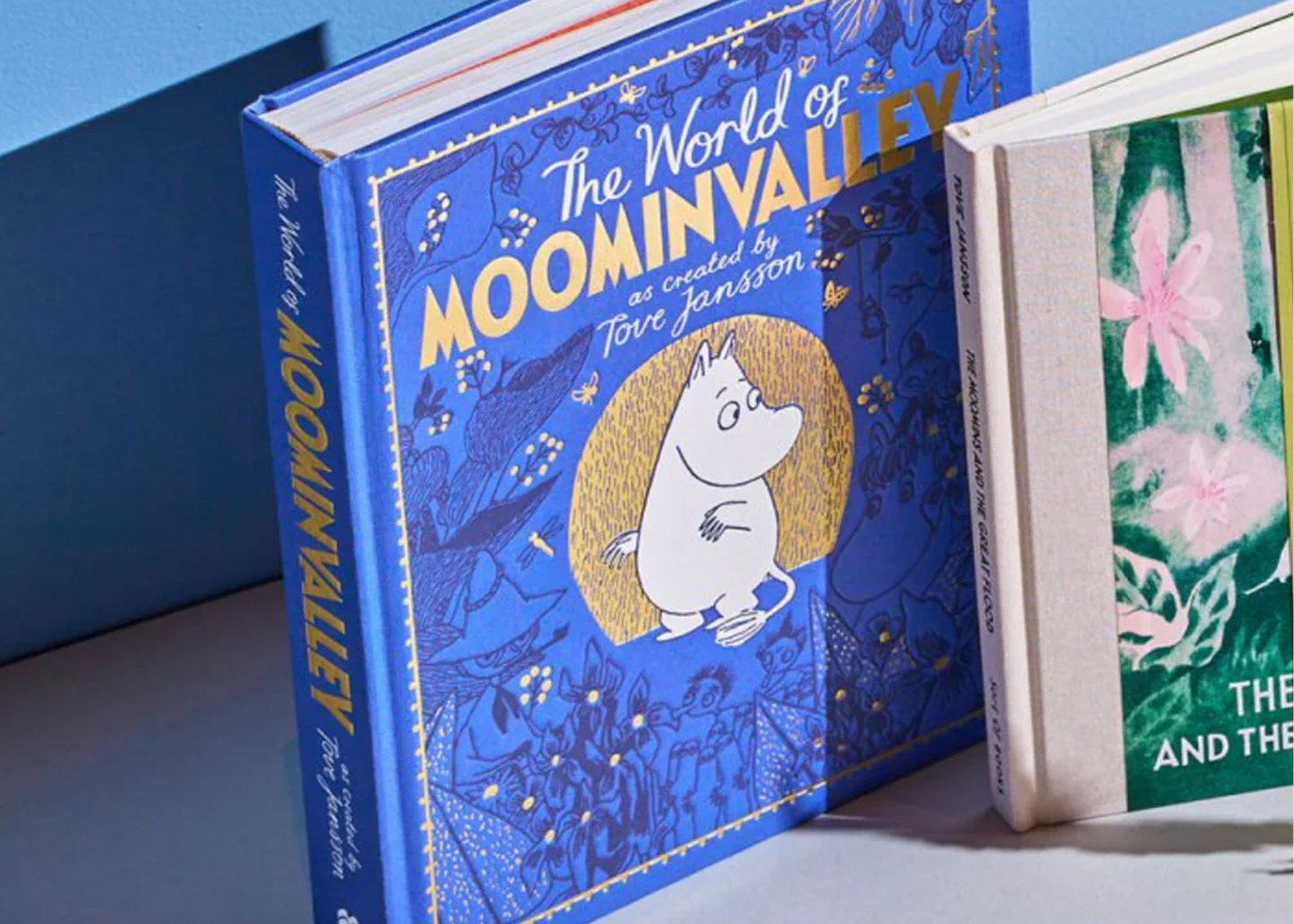 A close-up of a children's hardcover book titled "The World of Moominvalley" with a bright blue cover, gold lettering, and a central illustration of a white character in a gold circle. Part of another book with a green floral cover is visible at the right edge against a blue background.