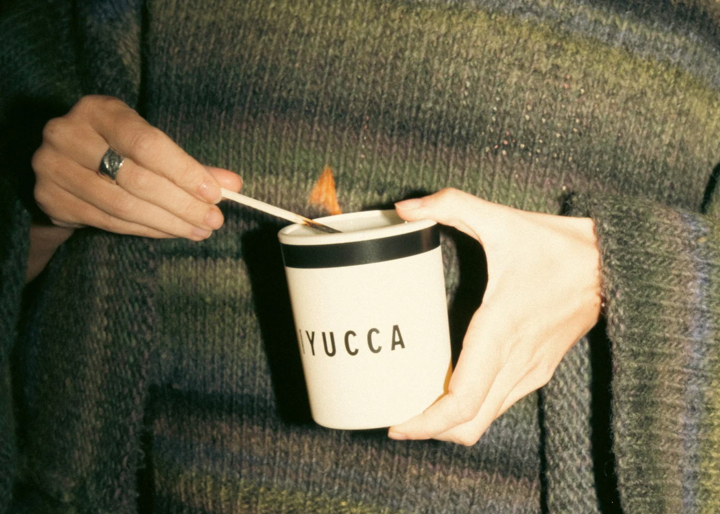 
                  
                    A person in a ribbed knit sweater holds a white candle jar with a black rim and the word 'YUCCA' printed on the front, while lighting it with a match. The background is the dark, cozy knit sweater.
                  
                