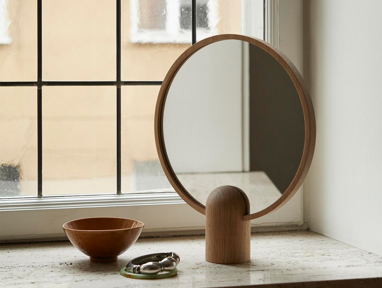 
                  
                    The round handheld large Aino mirror by Skagerak and Fritz Hansen in front of a window on a ledge
                  
                