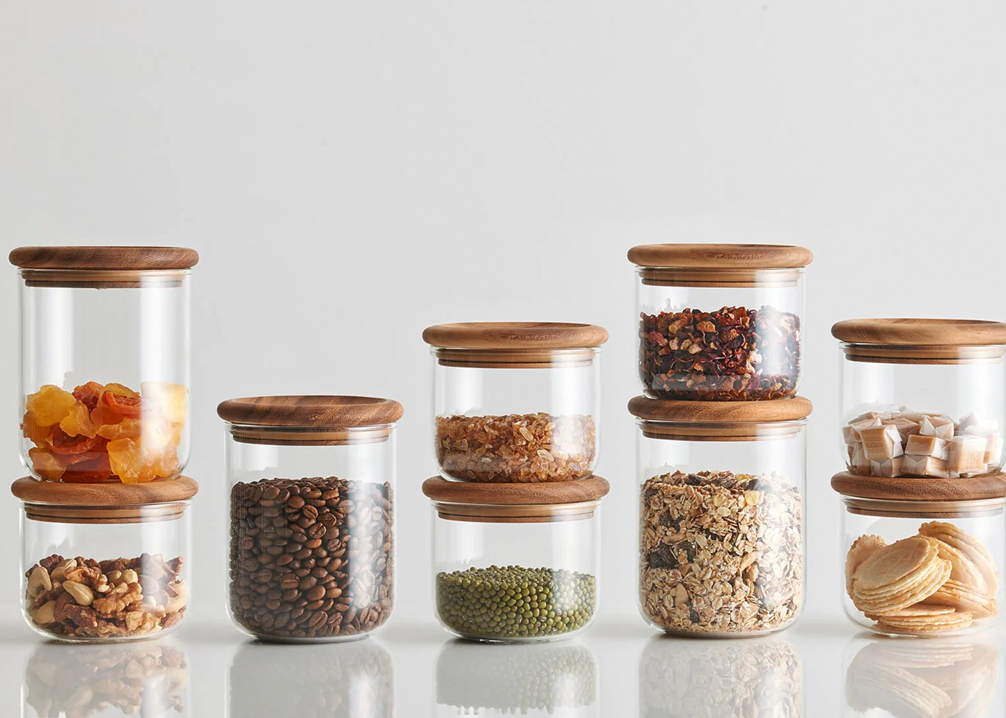 A collection of BAUM NEU clear glass canister with a wooden round lid by KINTO all each filled with spices and kitchen goods.