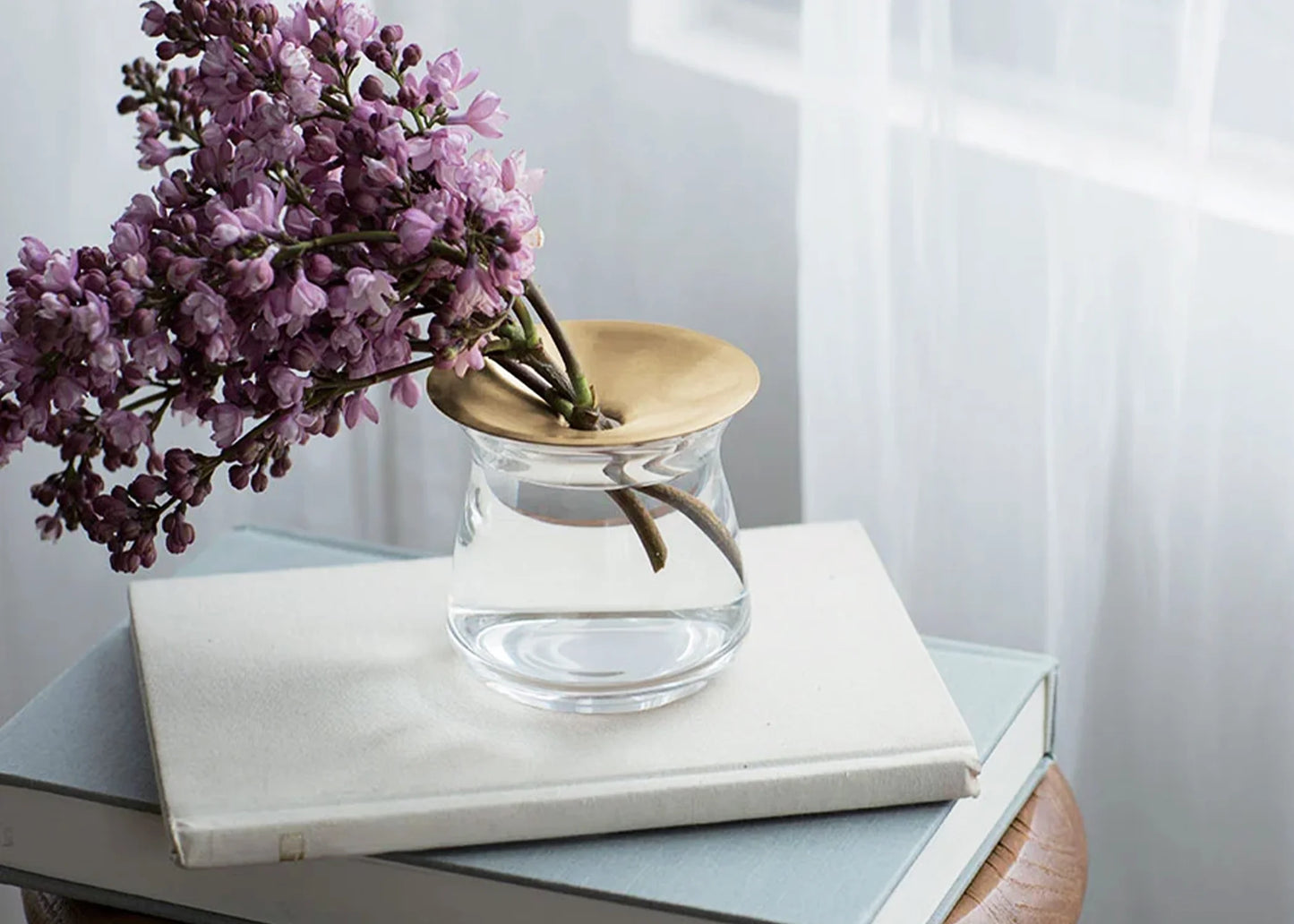 
                  
                    The small glass LUNA vase with gold lid holding a bunch of purple flowers on a desk with books.
                  
                