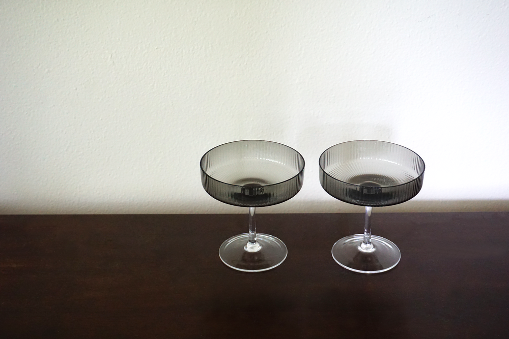 
                  
                    Grey smoked ripple champagne glasses by Ferm Living on a wooden table surface
                  
                